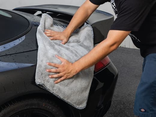 Person drying the rear of a black car with a large, gray microfiber towel, focusing on removing water after a wash